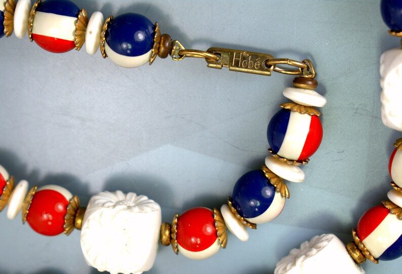 May include: A vintage Hobe necklace with red, white, and blue beads. The beads are round and have a gold trim. The necklace has a clasp with the Hobe logo.