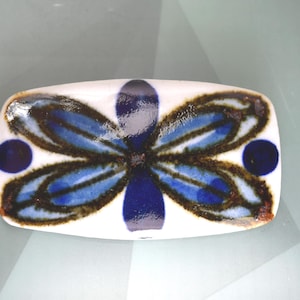 May include: A rectangular ceramic dish with a glossy white finish, featuring a hand-painted floral motif in deep blue and dark brown. The design consists of a central stylized flower with elongated petals and two solid blue circles on the sides.