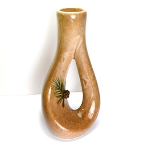 May include: A tan ceramic vase with a unique teardrop shape and a large opening. The vase features a painted pine branch and pinecone design. The vase is approximately 10 inches tall.