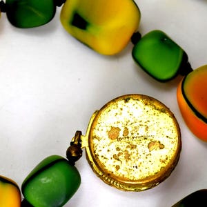 Op de afbeelding: Close-up van een vintage ketting met grote, kleurrijke kralen in geel, groen en oranje. De kralen zijn geregen met een goudkleurige sluiting. De ketting is waarschijnlijk een statement-sieraad.