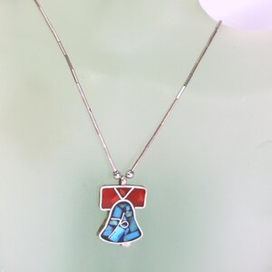 Peut inclure: Un collier en argent avec un pendentif en forme de cloche. La cloche est en argent et est incrust&eacute;e de turquoise et de pierres rouges.