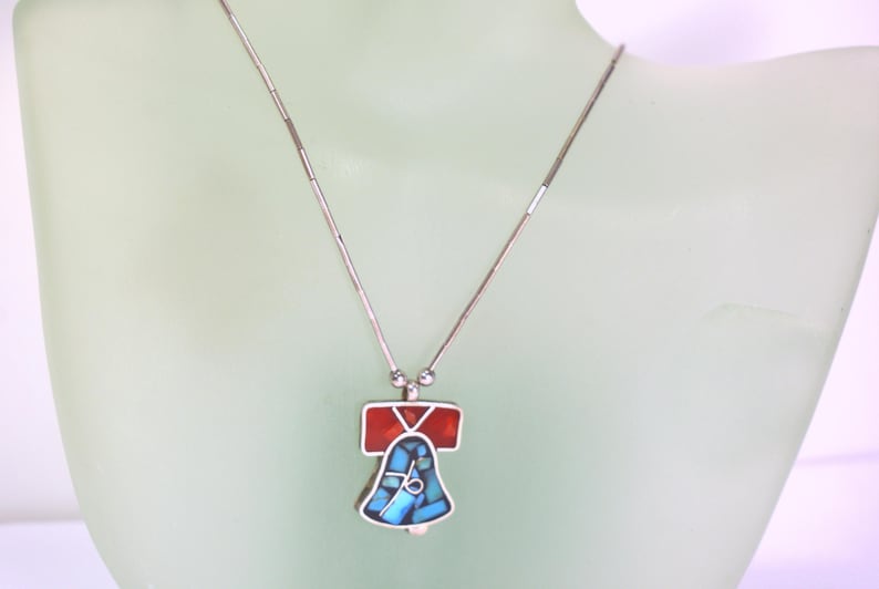 Peut inclure: Un collier en argent avec un pendentif en forme de cloche. La cloche est en argent et est incrust&eacute;e de turquoise et de pierres rouges.