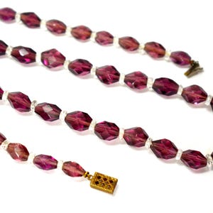 May include: A vintage necklace with faceted, deep purple beads and clear spacer beads. The necklace has a gold-toned clasp. The beads are arranged in a symmetrical pattern. The necklace is displayed against a white background.