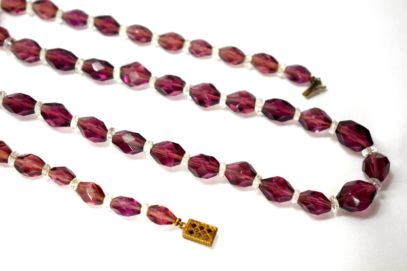 May include: A vintage necklace with faceted, deep purple beads and clear spacer beads. The necklace has a gold-toned clasp. The beads are arranged in a symmetrical pattern. The necklace is displayed against a white background.