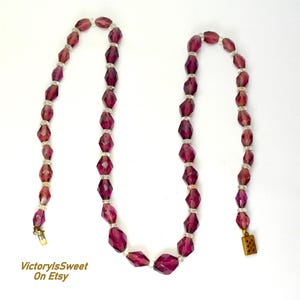May include: A vintage necklace with faceted, deep purple beads and smaller, clear beads. The necklace has a gold-toned clasp and is set against a white background. The beads are geometric and strung together. The necklace is a piece of jewellery.