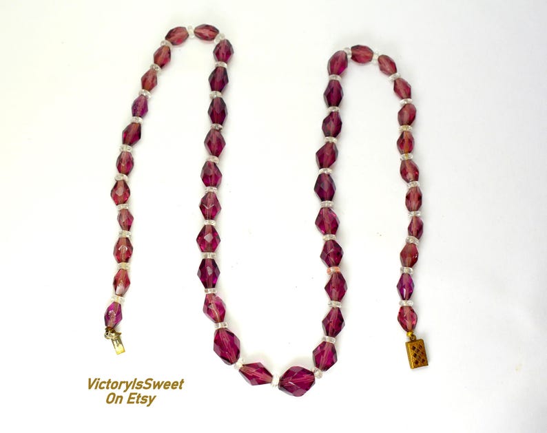 May include: A vintage necklace with faceted, deep purple beads and smaller, clear beads. The necklace has a gold-toned clasp and is set against a white background. The beads are geometric and strung together. The necklace is a piece of jewellery.