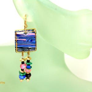 May include: Gold-toned earrings with a square, marbled design in blue, pink, and black, framed in gold. The earrings dangle with colorful beads in pink, green, black, and blue, creating a vibrant accessory.