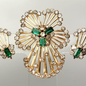 May include: A gold-tone brooch and earrings set with clear rhinestones and emerald green stones. The brooch is a large, abstract flower shape with the stones set in a radiating pattern. The earrings are smaller, fan-shaped designs with a single emerald green stone at the centre.