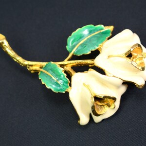May include: A gold-tone brooch featuring two white enamel roses with gold accents, set on a green enamel stem with leaves.