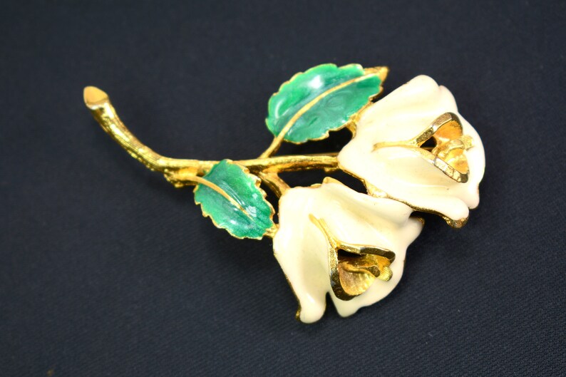 May include: A gold-tone brooch featuring two white enamel roses with gold accents, set on a green enamel stem with leaves.