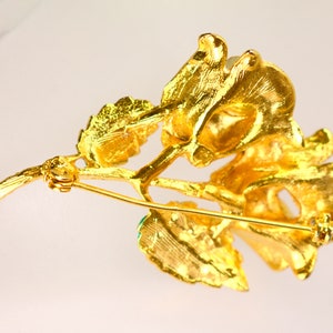 May include: A gold-toned brooch in the shape of a flower with green gemstone accents. The brooch has a pin and catch closure.