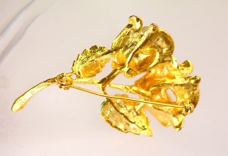 May include: A gold-toned brooch in the shape of a flower with green gemstone accents. The brooch has a pin and catch closure.