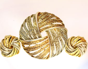 Op de afbeelding: Een sieradenset van goudkleurig metaal bestaande uit een grote ronde broche en twee bijpassende ronde oorbellen. Elk stuk heeft een gestructureerd, wervelend lintontwerp met afwisselend gepolijste en geborstelde metalen afwerkingen.