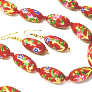 May include: A red beaded necklace and matching earrings set. The oval beads are painted with floral designs in pink, blue, and yellow. The jewelry has gold-coloured hooks and connectors.
