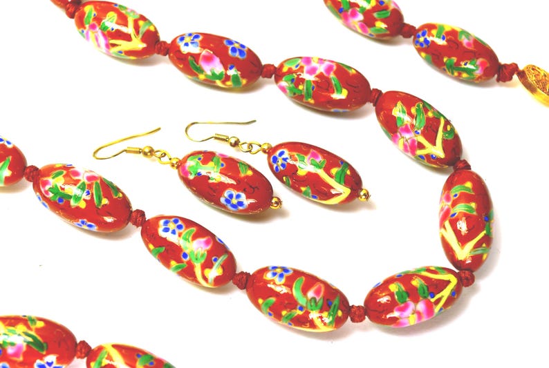 May include: A red beaded necklace and matching earrings set. The oval beads are painted with floral designs in pink, blue, and yellow. The jewelry has gold-coloured hooks and connectors.