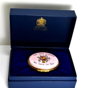 May include: A navy blue box with a gold emblem on the lid, containing a pink oval trinket box. The trinket box has a gold rim and features a floral design with the text "Let this Convey My Love to You."