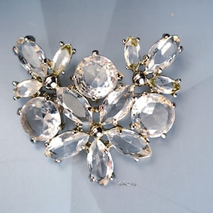 May include: A silver-toned brooch featuring a cluster of clear, faceted, marquise-cut and round-cut crystals arranged in a symmetrical, fan-like pattern. The metal setting holds the stones securely, creating a reflective and multi-faceted appearance.