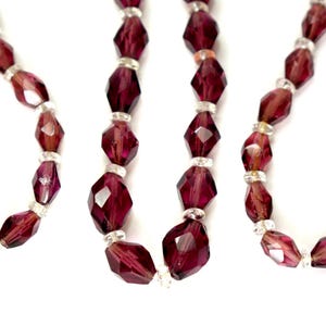 May include: Close-up of several strands of faceted, deep burgundy beads. The beads are separated by small, clear, round beads. The necklaces have a vintage aesthetic and are displayed against a white background.