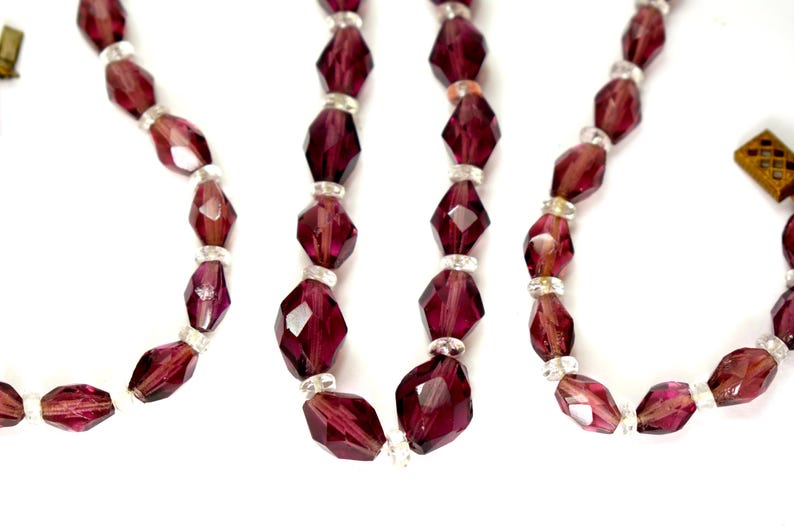 May include: Close-up of several strands of faceted, deep burgundy beads. The beads are separated by small, clear, round beads. The necklaces have a vintage aesthetic and are displayed against a white background.