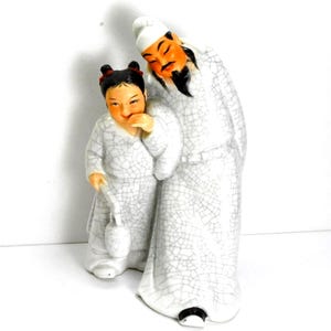 May include: A ceramic figurine depicting two figures in traditional Asian attire. The figures are predominantly white with a crackle glaze, and feature orange and black facial details. One figure is taller, leaning over the smaller figure.