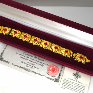 May include: A gold-toned bracelet with red gemstone accents, presented in a burgundy velvet-lined box. The bracelet features a repeating floral design. Accompanying documents are visible, including a certificate and a description.