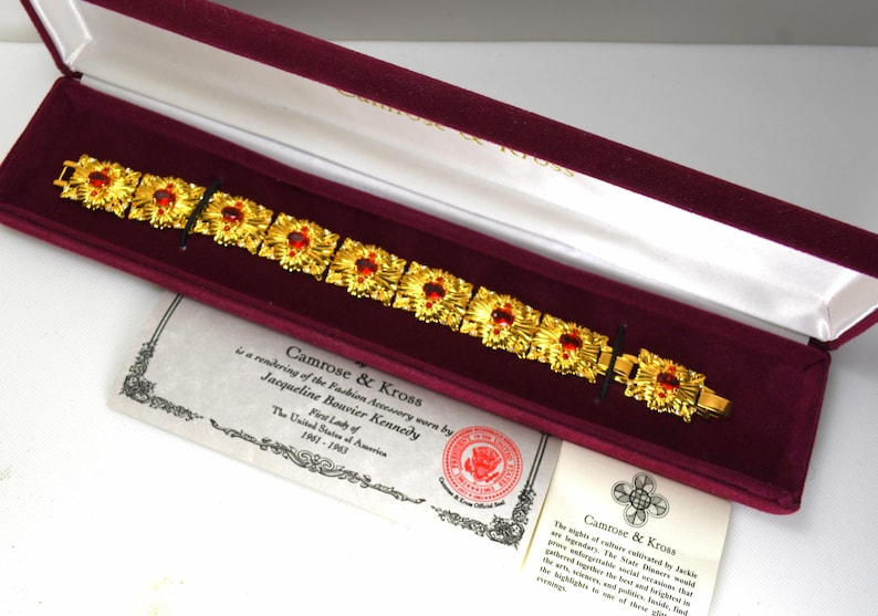 May include: A gold-toned bracelet with red gemstone accents, presented in a burgundy velvet-lined box. The bracelet features a repeating floral design. Accompanying documents are visible, including a certificate and a description.