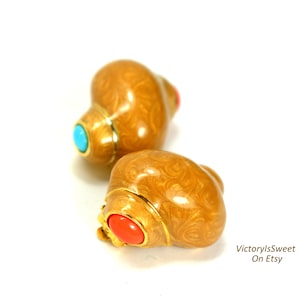 Può includere: Due orecchini dorati a forma di conchiglia con un motivo a spirale. Ogni orecchino presenta un bordo dorato e una piccola gemma rotonda, turchese o corallo. Su sfondo bianco.