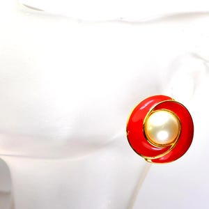 以下が含まれることがあります： 赤と金のクリップオンイヤリング。中央に大きく丸いクリーム色のイミテーションパール。イヤリングは金色の縁取りと鮮やかな赤のエナメル仕上げ。デザインはスパイラル状です。