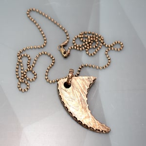 May include: A gold chain necklace with a bronze pendant shaped like a crescent moon. The pendant has a textured surface.