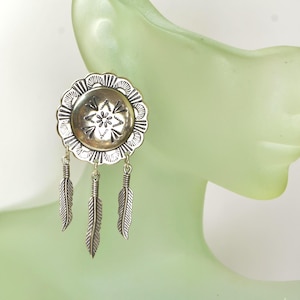May include: A silver-toned earring featuring a circular concho-style disc with scalloped edges and stamped geometric patterns. Three small, detailed metal feather charms hang from the bottom of the disc by thin jump rings, creating a dangling effect.