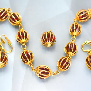 Könnte beinhalten: Ein goldfarbenes Schmuckset mit Halskette, Ohrringen und einer einzelnen Perle. Halskette und Ohrringe haben ein sich wiederholendes Muster aus roten, facettierten Perlen, die in goldenen Filigran-Käfigen eingeschlossen sind. Die Ohrringe sind Clip-On-Stil.