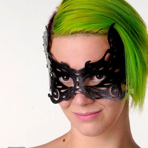 May include: A woman with bright green hair wears a black lace masquerade mask. The mask covers her eyes and has intricate details.