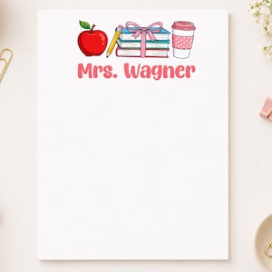 May include: White notepad with a colorful illustration of an apple, pencil, books, and coffee cup. The text "Mrs. Wagner" is printed in pink. Gold paper clips and pink binder clips are scattered around the notepad.