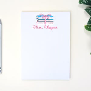 May include: White notepad with a stack of books illustration tied with a pink bow. The notepad has the name "Mrs. Wagner" printed in pink script. A silver pencil is on the left side of the notepad.