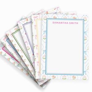 May include: A stack of rectangular stationery note cards featuring a repeating pattern of small pink flowers and light blue bows. Each card has a solid colored border and the name SAMANTHA SMITH printed in pink capital letters at the top center.