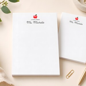 two blank white notepads with personalized names on them, along with a gold pen and a gold paper clip, all placed on a light-colored surface.