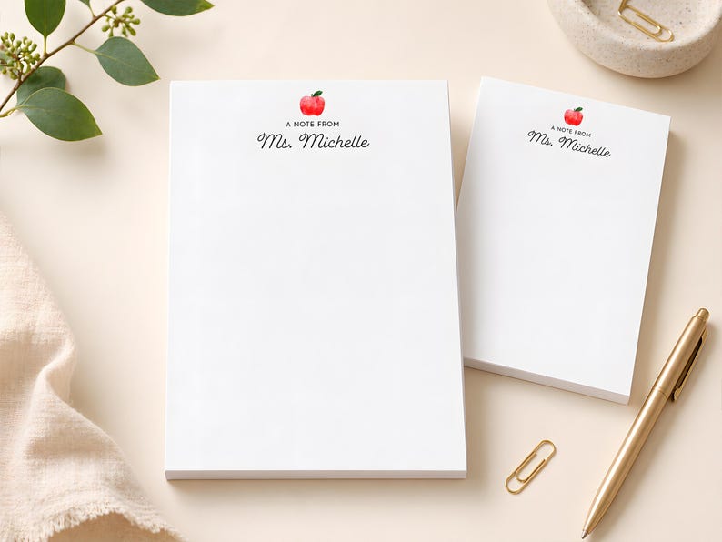 two blank white notepads with personalized names on them, along with a gold pen and a gold paper clip, all placed on a light-colored surface.