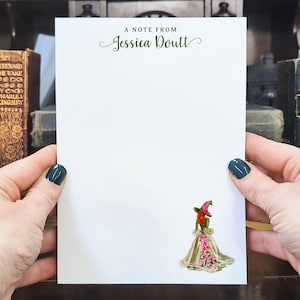 May include: A white notepad with the text "A Note From Jessica Doutt" in green script. A watercolor illustration of a witch in a dress and hat is at the bottom. The notepad is held by two hands with dark green nail polish.