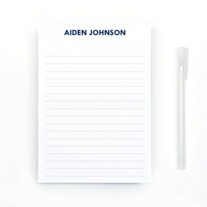 May include: A white notepad with blue lined paper and the name "AIDEN JOHNSON" printed at the top. A clear pen is lying next to the notepad.