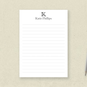 May include: A white lined notepad with the name "Katie Phillips" and a decorative "K" at the top. A gray pencil with a pink eraser rests on the right side of the notepad.