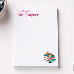 May include: A white rectangular notepad featuring the pink printed text a note from Mrs. Wagner at the top and a watercolor illustration of a stack of four colorful books topped with a small orange flower in the bottom right corner.