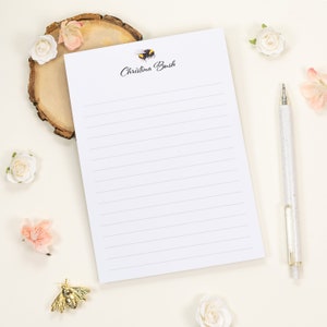 May include: A white lined notepad featuring a small illustration of a bumblebee and the printed name Christina Bush at the top. The notepad is accompanied by a speckled white pen, a circular wood slice, and several small white and pink flowers.