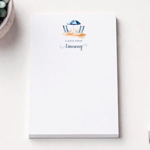 May include: A white rectangular notepad featuring a watercolor illustration of two blue and white striped beach chairs under a blue umbrella on orange sand. The text A NOTE FROM Tammy is printed in blue ink below the beach scene.
