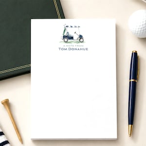 May include: A white notepad featuring a watercolor illustration of a blue golf cart at the top, followed by the printed text A NOTE FROM TOM DONAHUE in dark blue capital letters. The notepad is centered on a light surface with office items.