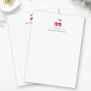 May include: A set of two white notepad stationery with a cherry design. The top notepad has the text "a note from LAUREN WILSON" and the bottom notepad has the text "MARI".