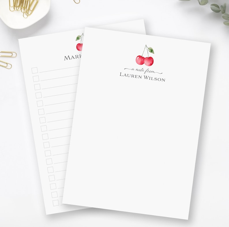 May include: A set of two white notepad stationery with a cherry design. The top notepad has the text "a note from LAUREN WILSON" and the bottom notepad has the text "MARI".