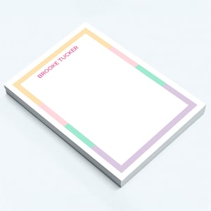 May include: A stack of white notepads with a colorful border. The border is composed of pastel colors: yellow, pink, green, and purple. The text "BROOKE TUCKER" is printed in pink in the upper left corner.
