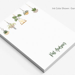 May include: White notepad with a green ink inscription "Pat Andrews" at the bottom. The top of the notepad features a line of five hanging potted plants with green leaves and brown pots.