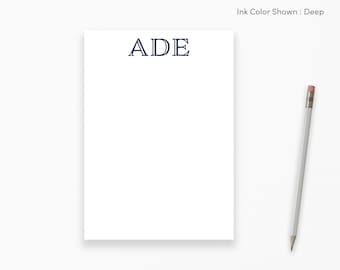 May include: A white notepad with the letters "ADE" printed in navy blue at the top. A pencil is shown to the right of the notepad.