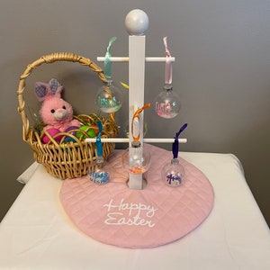 May include: A white wooden stand with four clear glass ornaments hanging from it. The ornaments are filled with colorful confetti and have the words "Happy Easter" written on them. The stand is sitting on a pink quilted mat with the words "Happy Easter" embroidered on it.